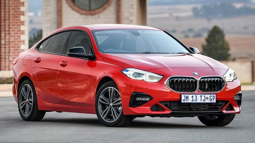 Vibrant red BMW 2 Series Gran Coupe parked on asphalt with a blurred brick building and hills in the background.