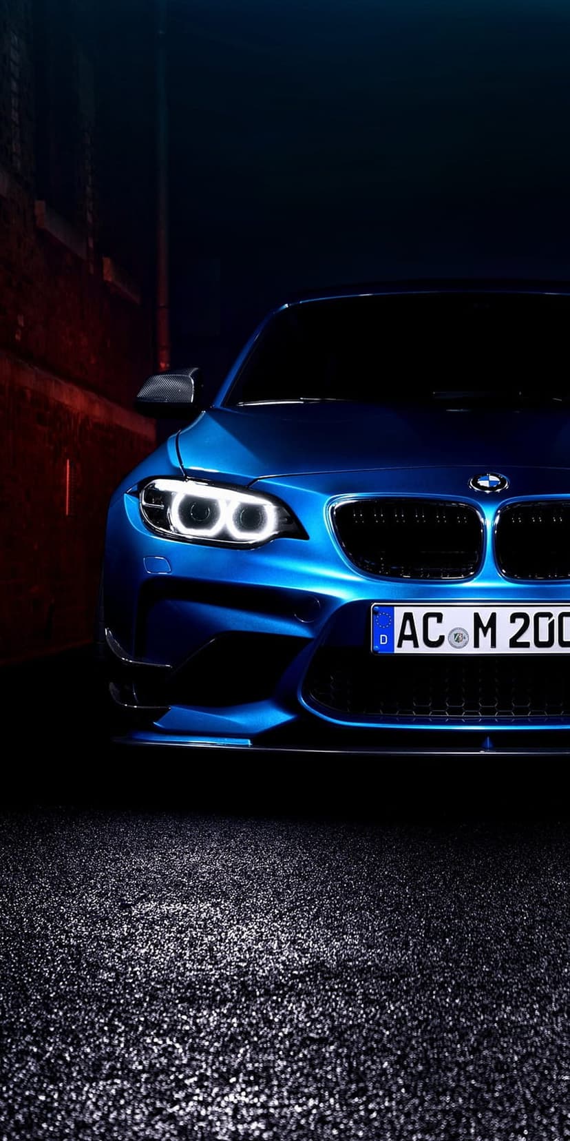 BMW M2 front view with illuminated headlights and blue paint on dark asphalt with a blurred red wall behind.