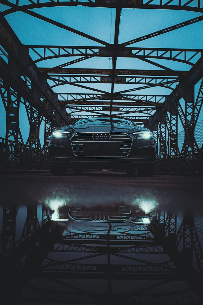 Black Audi car with glowing headlights parked on a wet surface reflects in a puddle under a steel bridge at night.