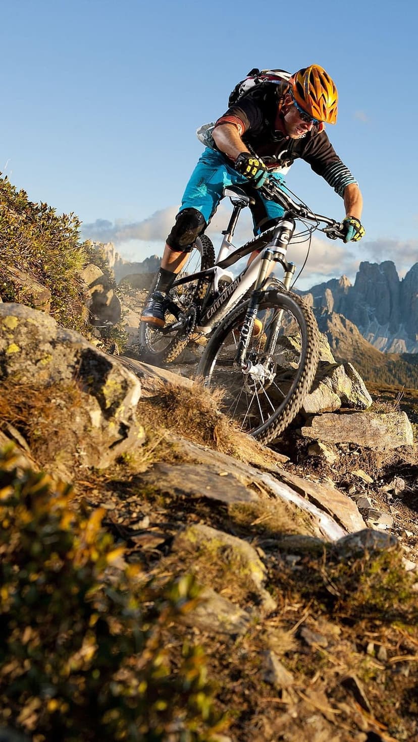 Mountain biker in full gear maneuvers a rocky slope with sharp gray peaks under a clear blue sky in the distance.
