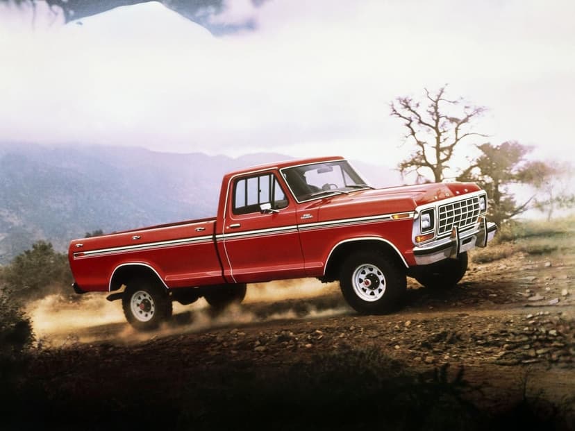 Red vintage Ford truck driving through dust on a mountain trail under a misty sky with a lone tree.