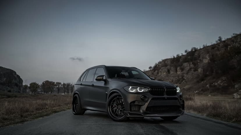 Matte black BMW X5 M with LED headlights on a rural road at dusk with rocky hills in the background.