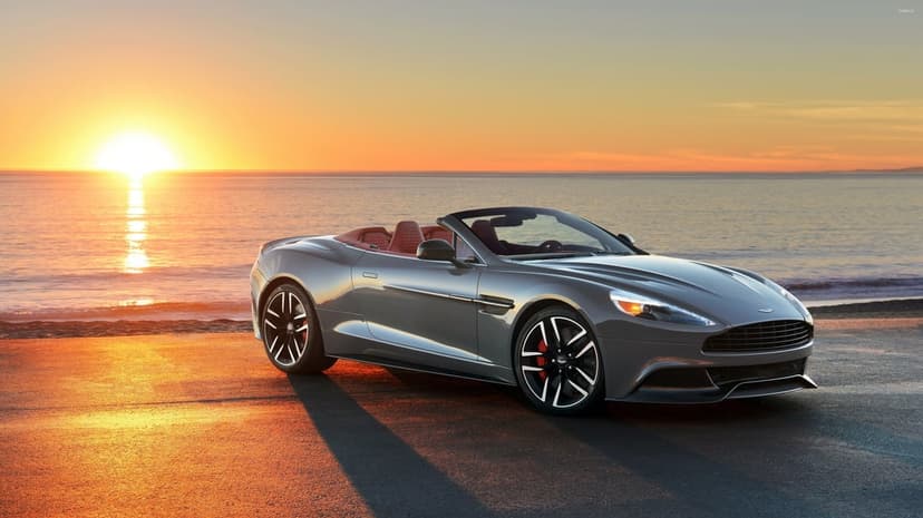 Grey convertible sports car parked on a beach road at sunset with golden light reflecting on the ocean.