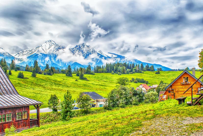 Weathered wooden cabins nestled in a lush green valley beneath jagged snow-dusted mountain peaks and grey clouds.