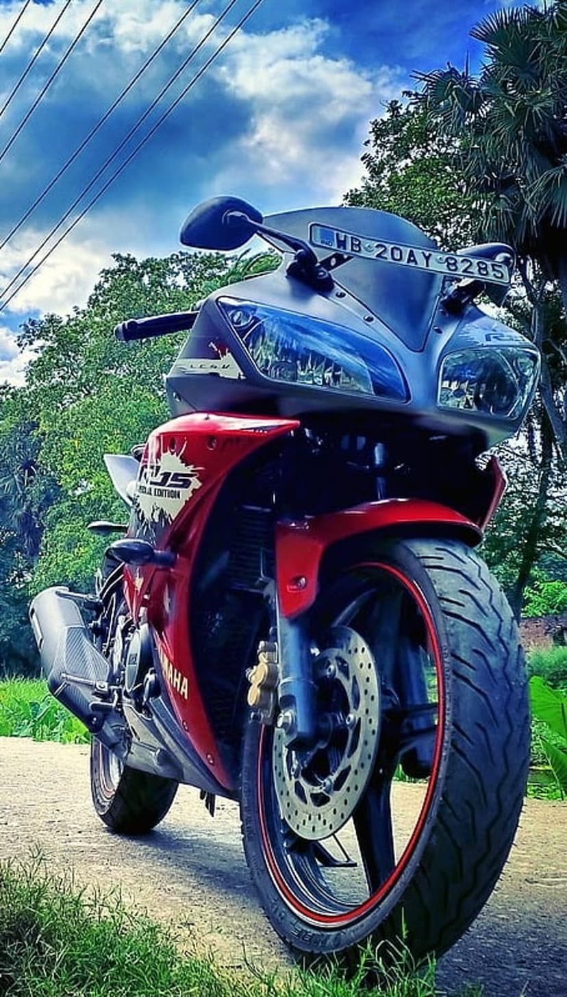 Red Yamaha R15 motorcycle parked on a dirt path with green grass under a dramatic blue sky with heavy clouds.