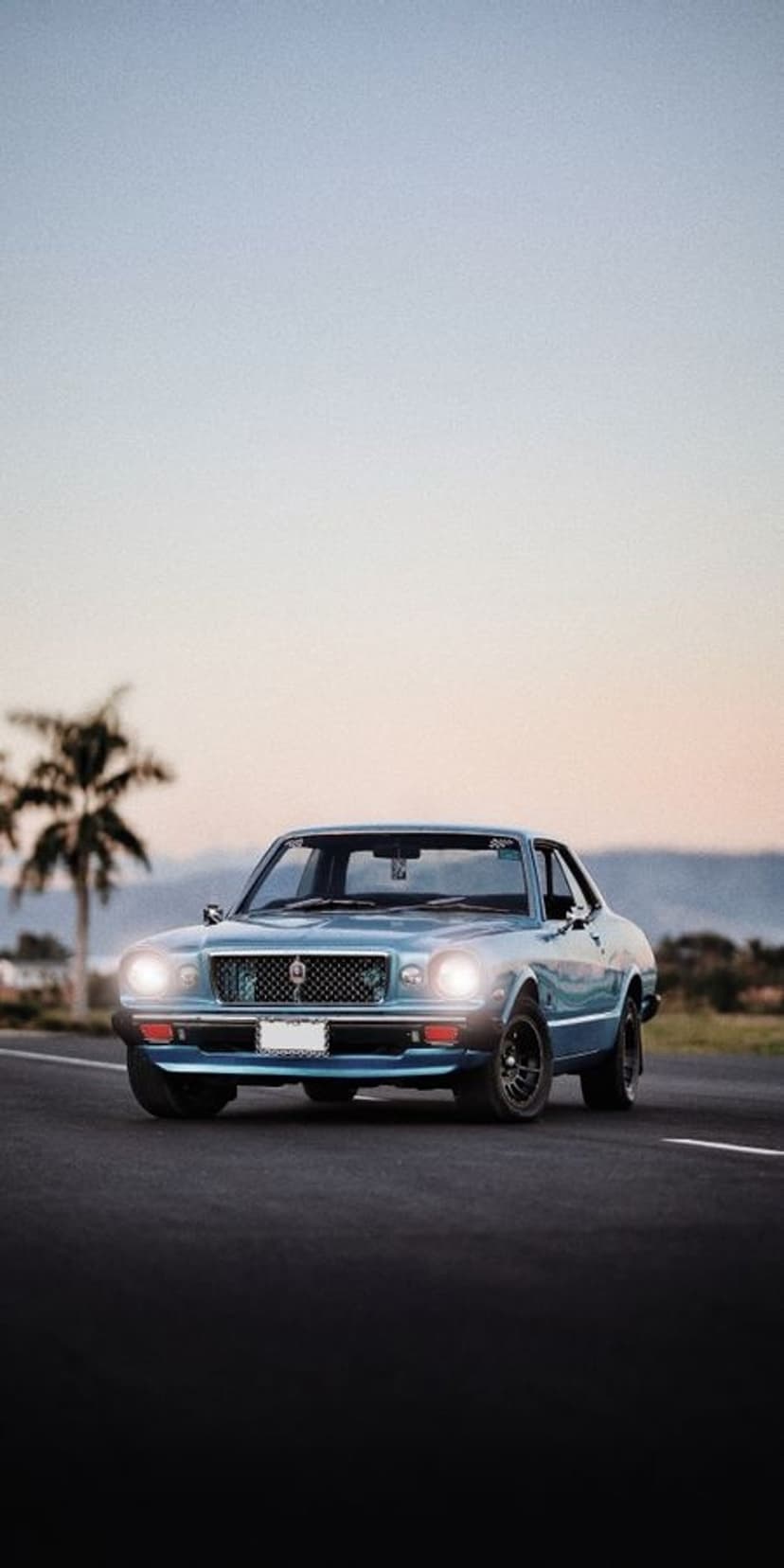 Blue vintage sedan drives along a coastal road past a palm tree under a soft orange and blue sunset sky.