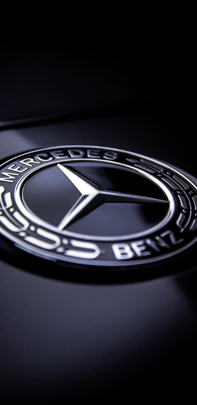 Mercedes-Benz silver emblem with sharp reflections on a glossy black surface with subtle overhead studio lighting.