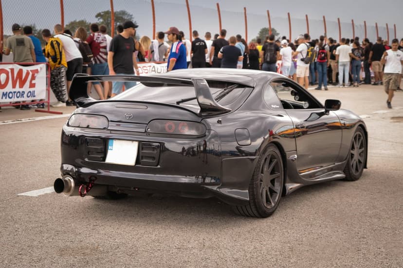 Black Toyota Supra MK4 with a large rear wing and aftermarket wheels parked at an outdoor car show with spectators.