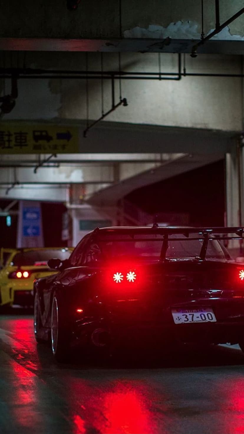 Mazda RX-7 with glowing red taillights sits on wet asphalt inside a dark tunnel next to another sports car.