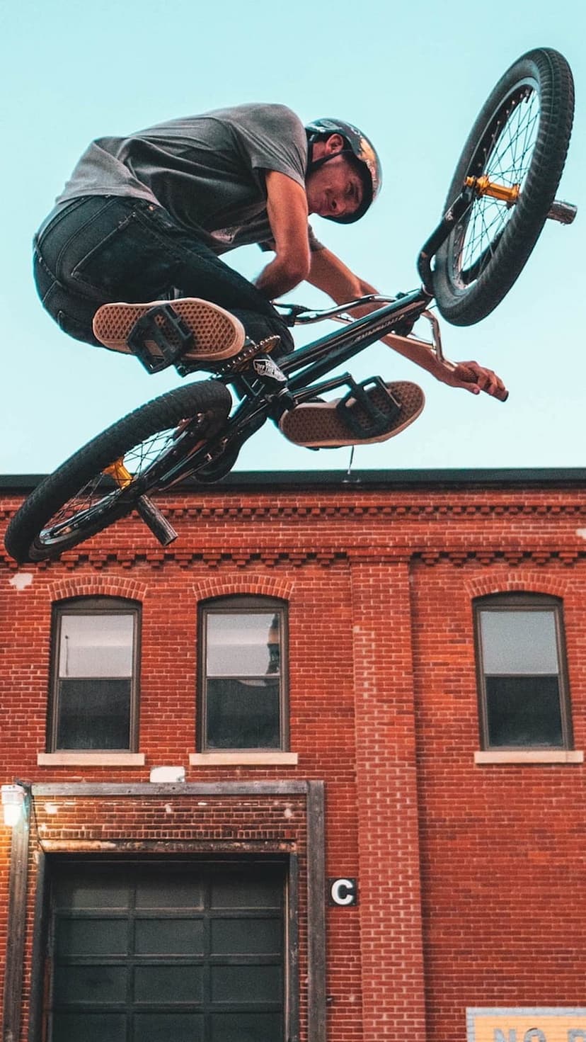 BMX rider performs a mid-air bicycle trick above a red brick warehouse under a clear blue sky during the day.