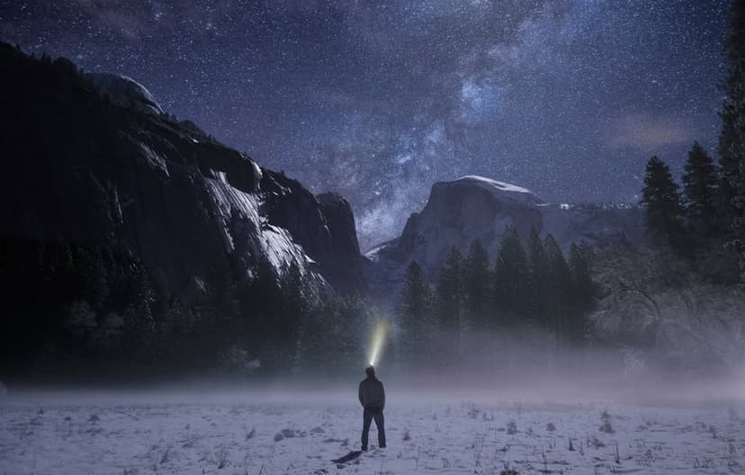 Lone hiker with a headlamp stands in snowy Yosemite Valley under a brilliant Milky Way and starry night sky.