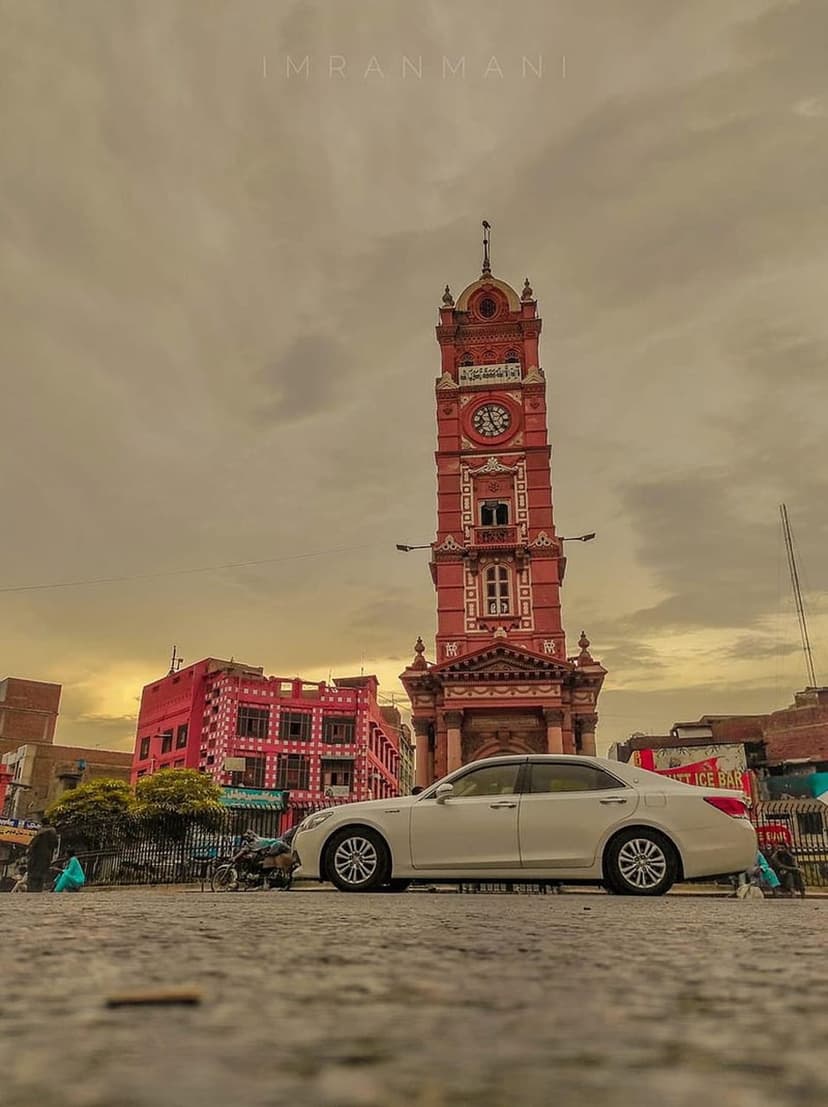 Red clock tower stands in an old city square next to a white sedan under a dramatic evening sky with clouds.
