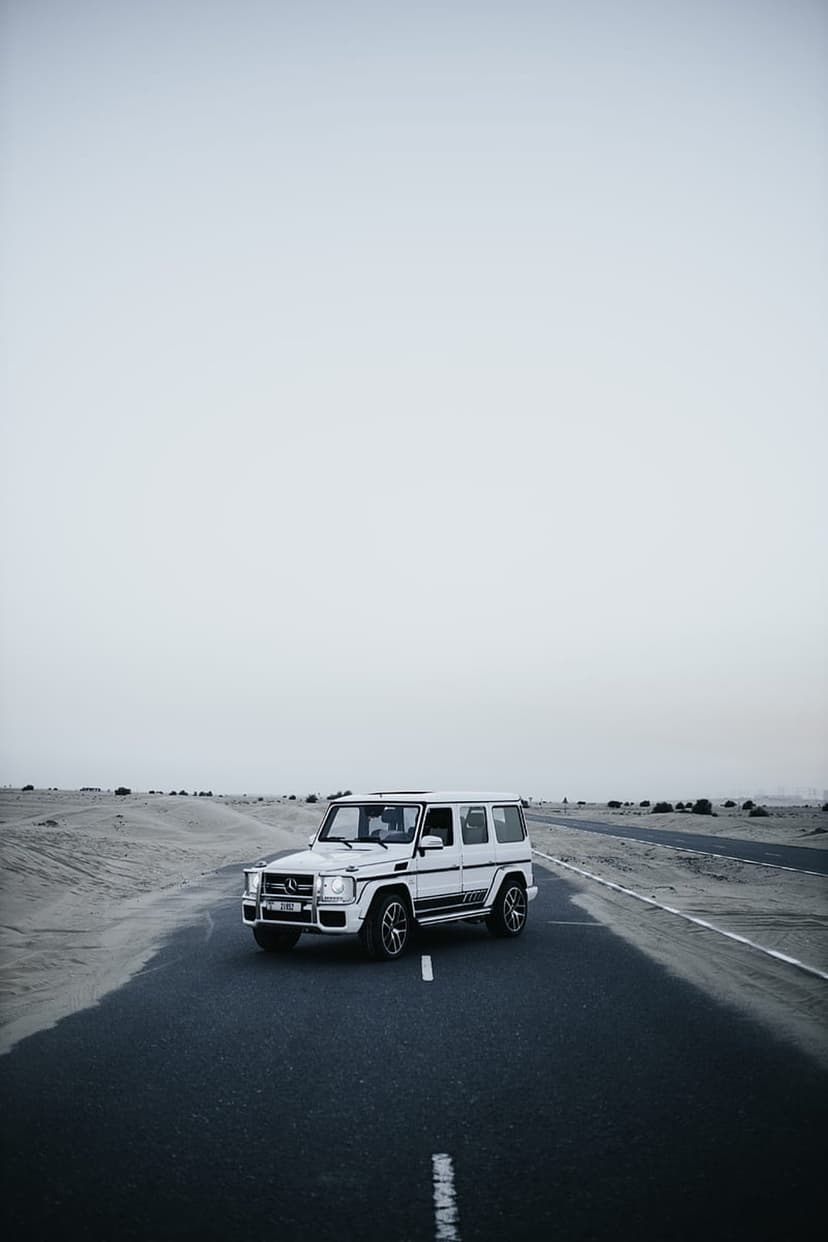 Mercedes G-Wagon driving on a straight desert highway under a pale overcast sky in a monochrome landscape scene.