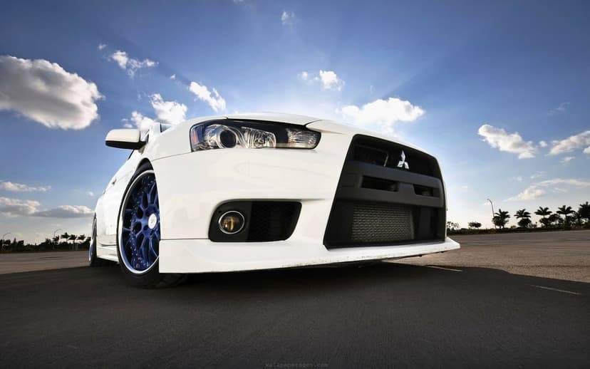 Low-angle view of a white Mitsubishi Lancer Evolution X with blue wheels under a bright blue sky with sun rays.
