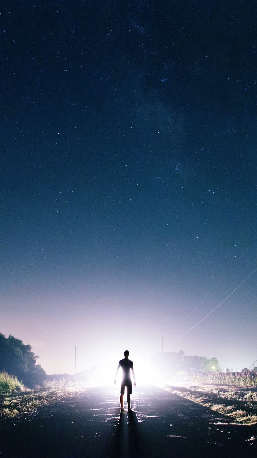 Lone person silhouetted by bright white light on a wet road at night under a clear starry sky with long shadows.