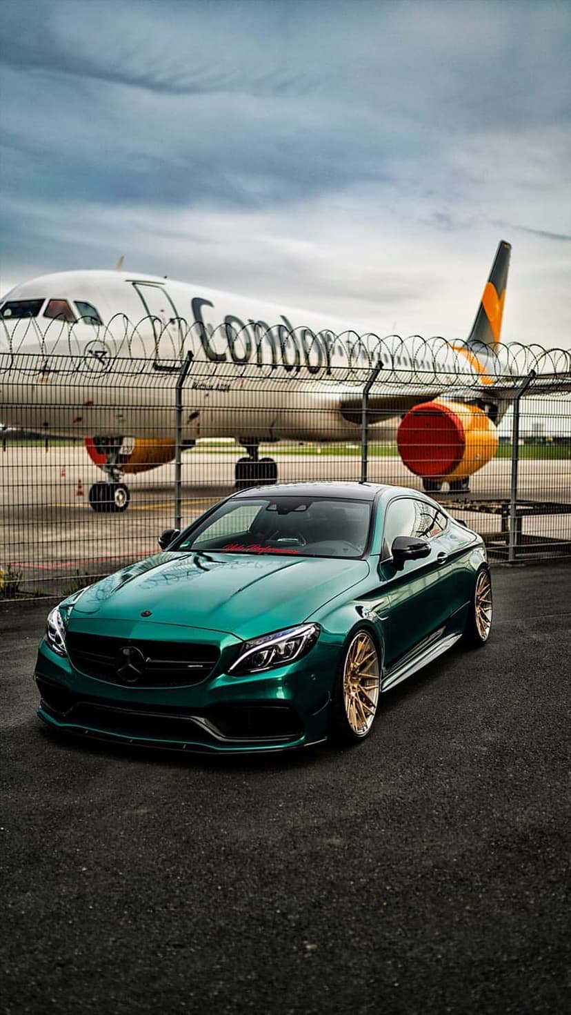 Emerald green Mercedes coupe with gold wheels parked on a runway in front of a striped Condor airplane.