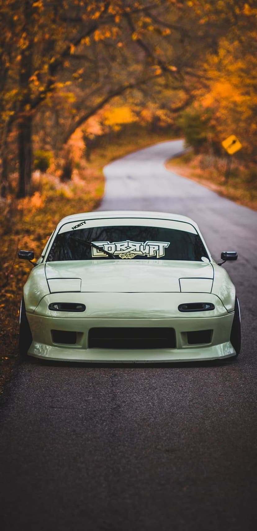 Light green Mazda Miata with pop-up headlights on a road lined with orange autumn trees and a yellow sign.