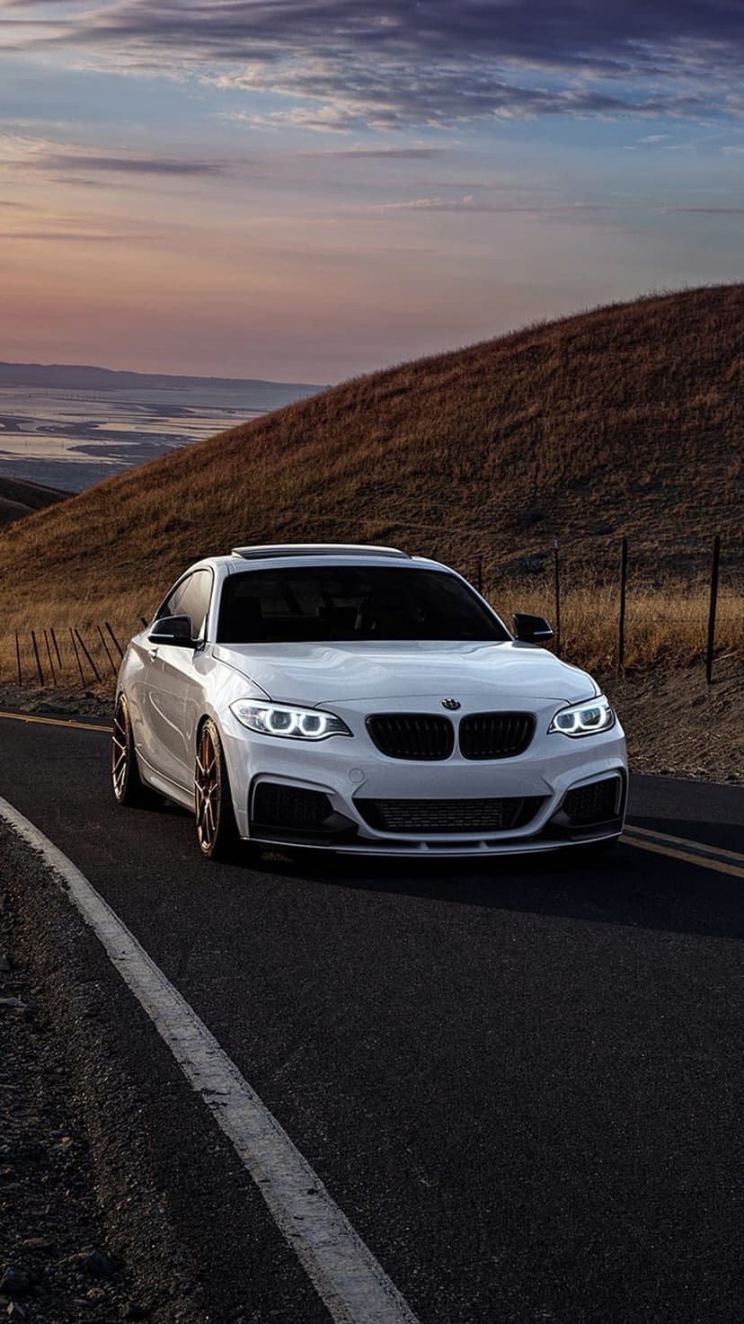 White BMW 2 Series Coupe with lit headlights cruises a winding mountain road under a pastel sunset sky.