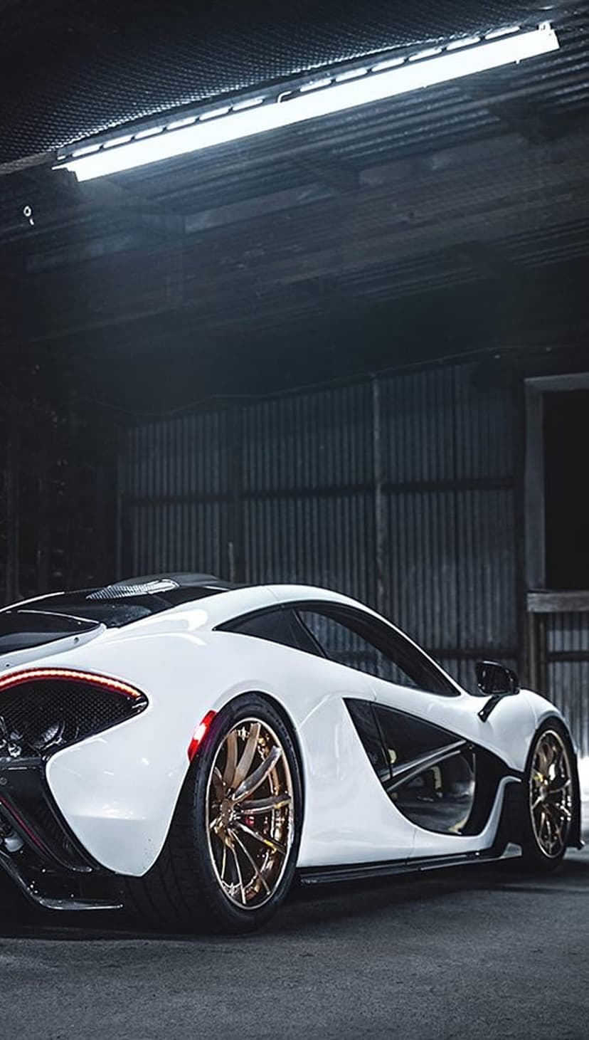 McLaren P1 supercar in white with gold alloy wheels parked against corrugated metal walls in a dim garage.