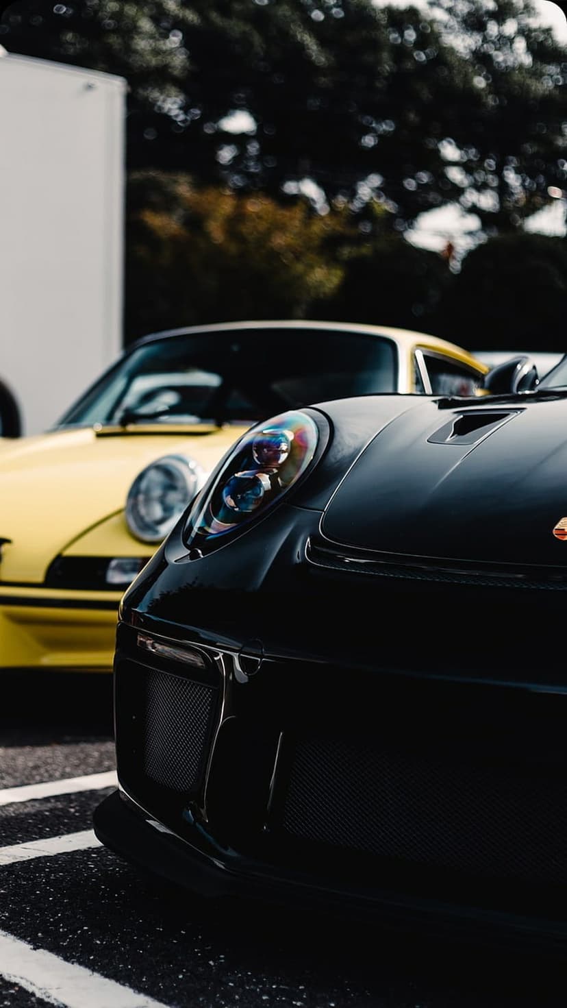 Black Porsche 911 headlight and front grille with a blurred yellow sports car fender in the background bokeh.