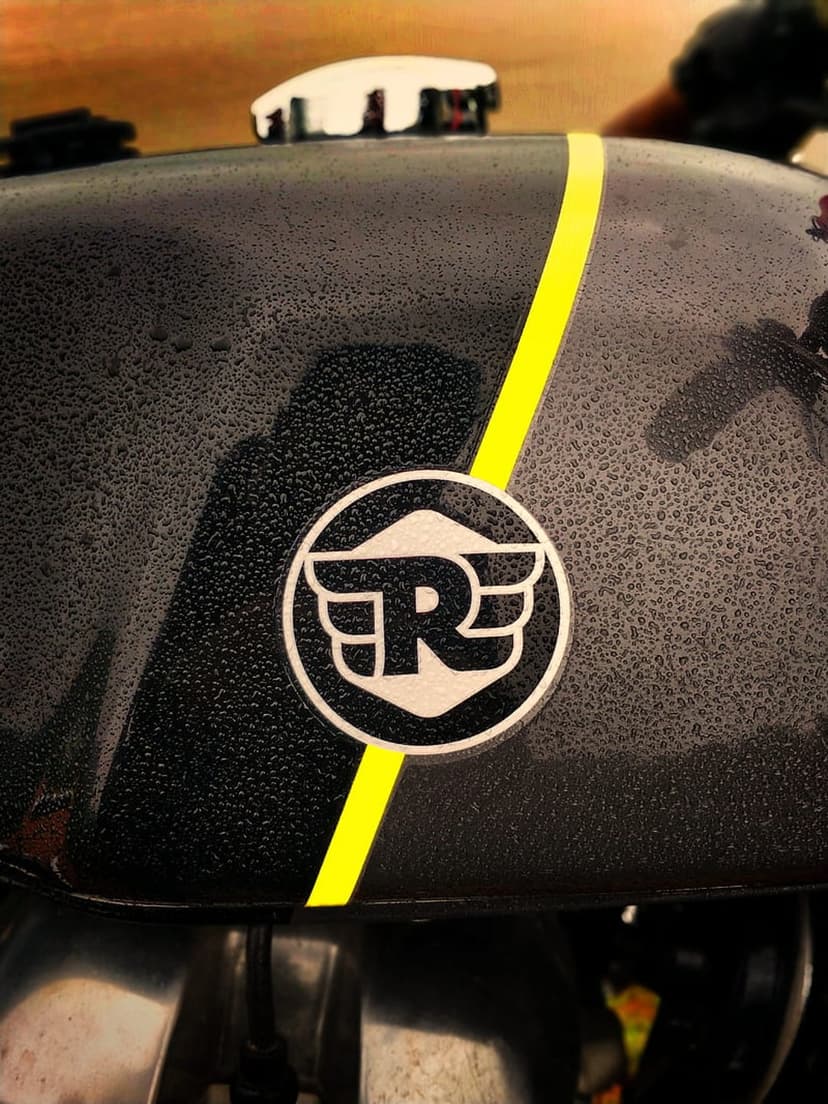 Royal Enfield winged emblem on a black fuel tank with a yellow stripe, chrome cap, and fresh water droplets.