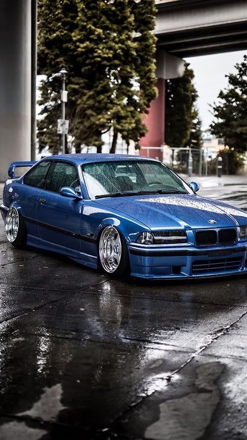 Blue BMW E36 M3 coupe with chrome wheels on wet pavement featuring reflections and an urban bridge background.