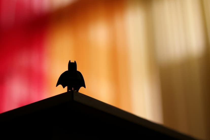 Batman silhouette stands on a dark building ledge against a blurred red and orange gradient sunset sky.