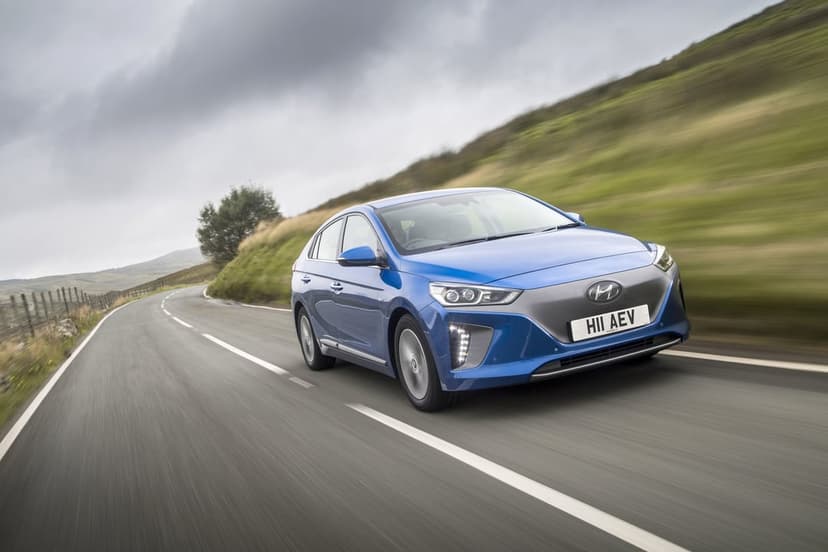 Blue Hyundai Ioniq Electric driving fast on a winding asphalt road through green hills under a cloudy sky.
