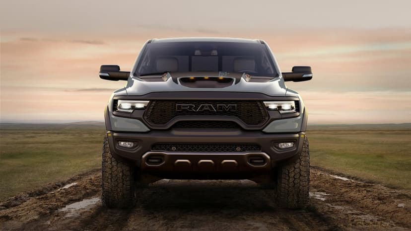 Front view of a grey Ram TRX truck with glowing headlights on a muddy path under a warm, glowing sunset sky.