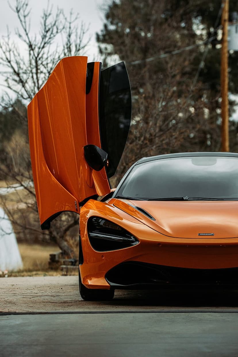 Orange McLaren 720S with open dihedral doors stands on a road against a blurred background of bare winter trees.