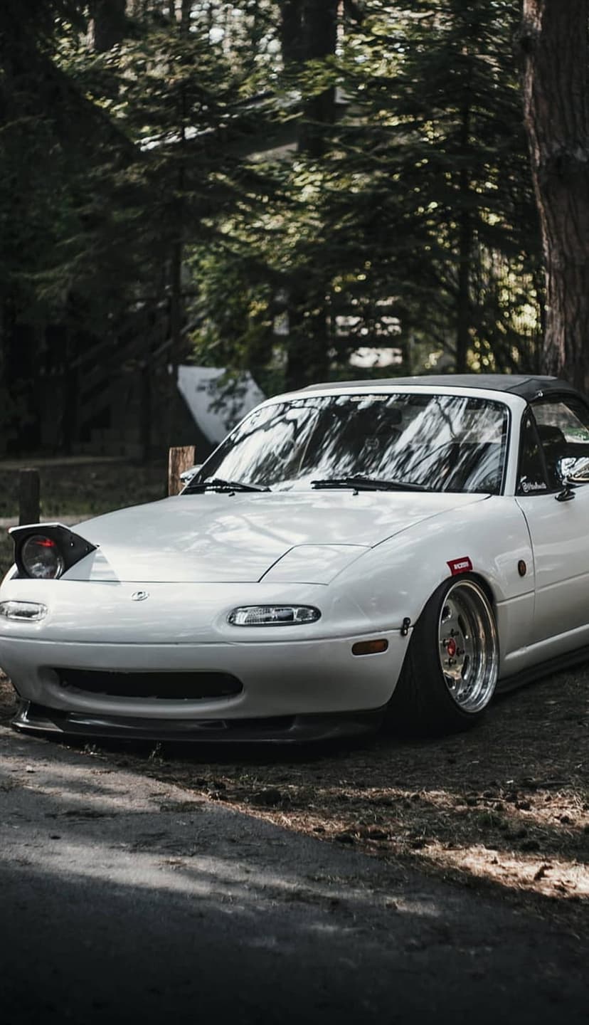 White Mazda Miata convertible with pop-up headlights parked on a dirt trail under a green forest canopy.