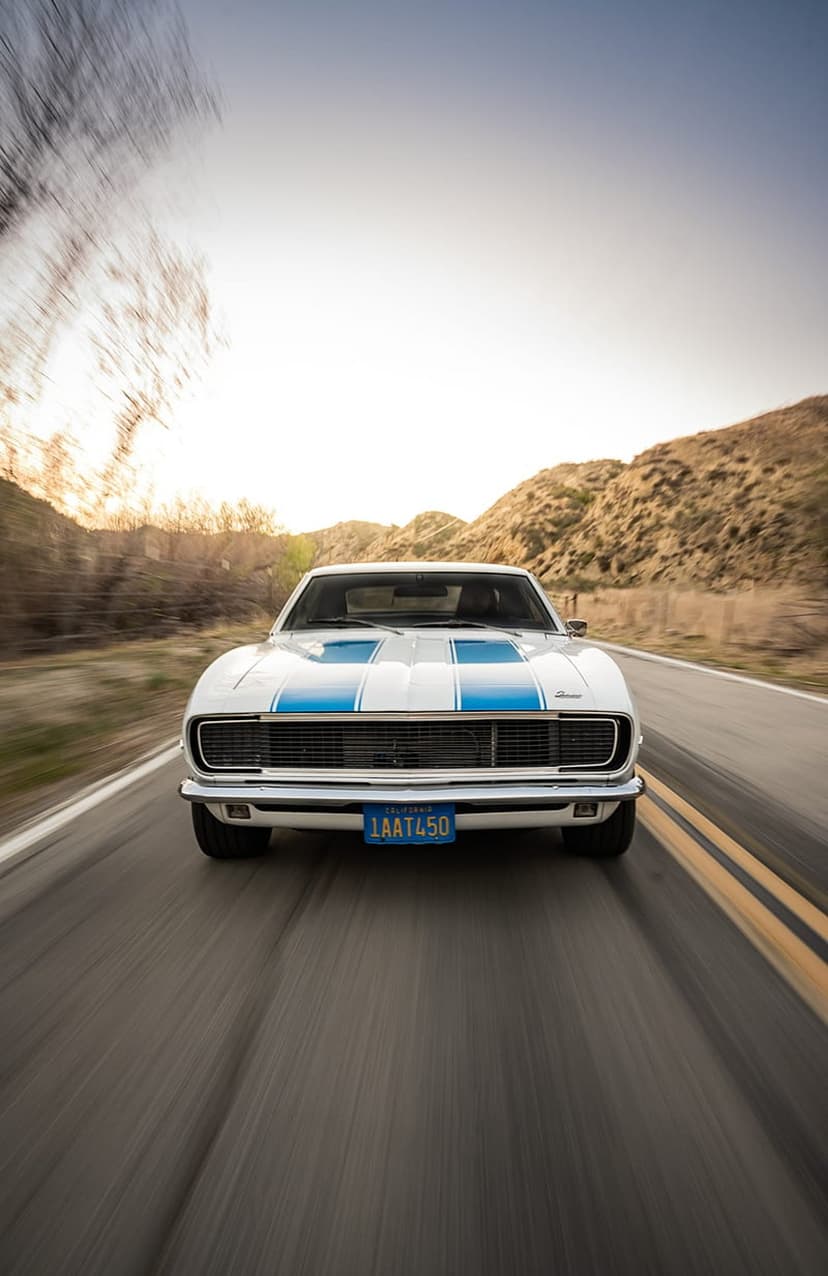 White vintage Camaro with blue stripes speeds down a desert highway under a warm golden sunset glow.