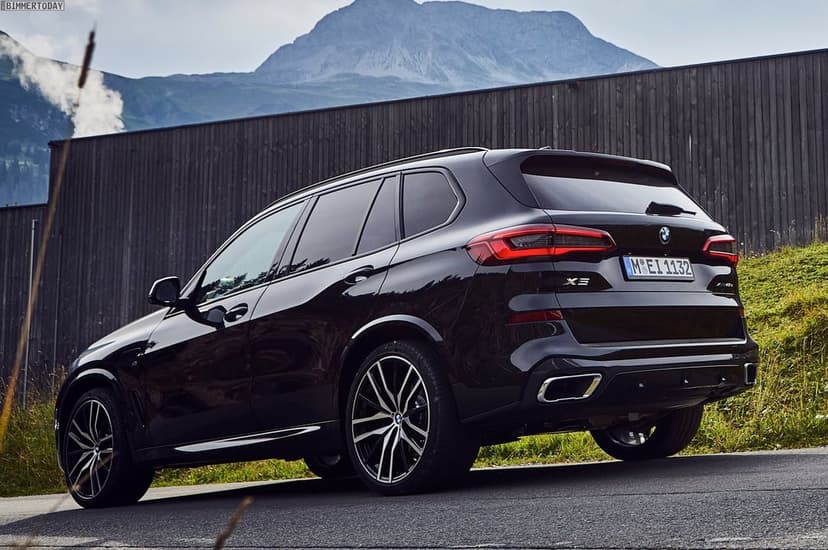 Black BMW X5 luxury SUV parked on an asphalt road with a large mountain peak and wooden fence in the background.