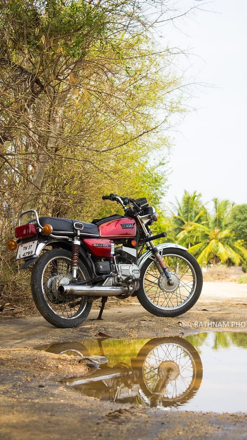 Red Yamaha RX 100 motorcycle reflecting in a water puddle on a dirt road surrounded by tropical palm trees.