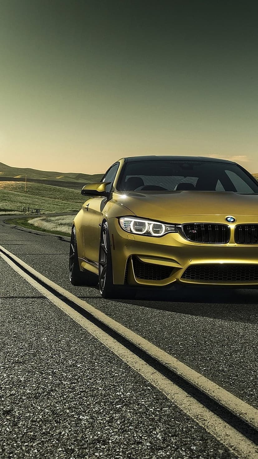 Golden BMW M4 sports car driving on a lonely asphalt road during a warm sunset with rolling hills in the background.