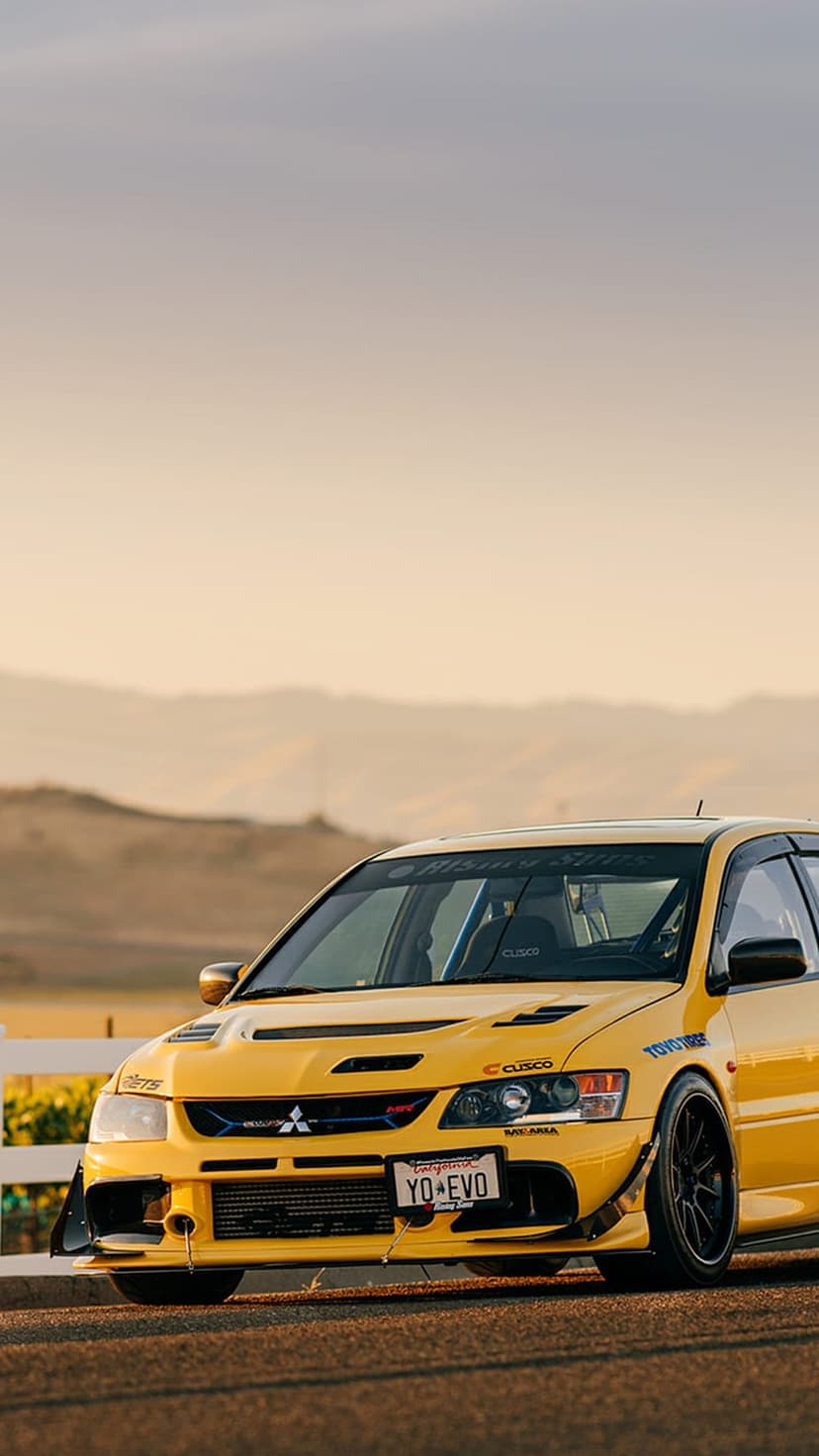 Yellow Mitsubishi Evo with racing decals parked on a mountain road during a hazy sunset with golden light.
