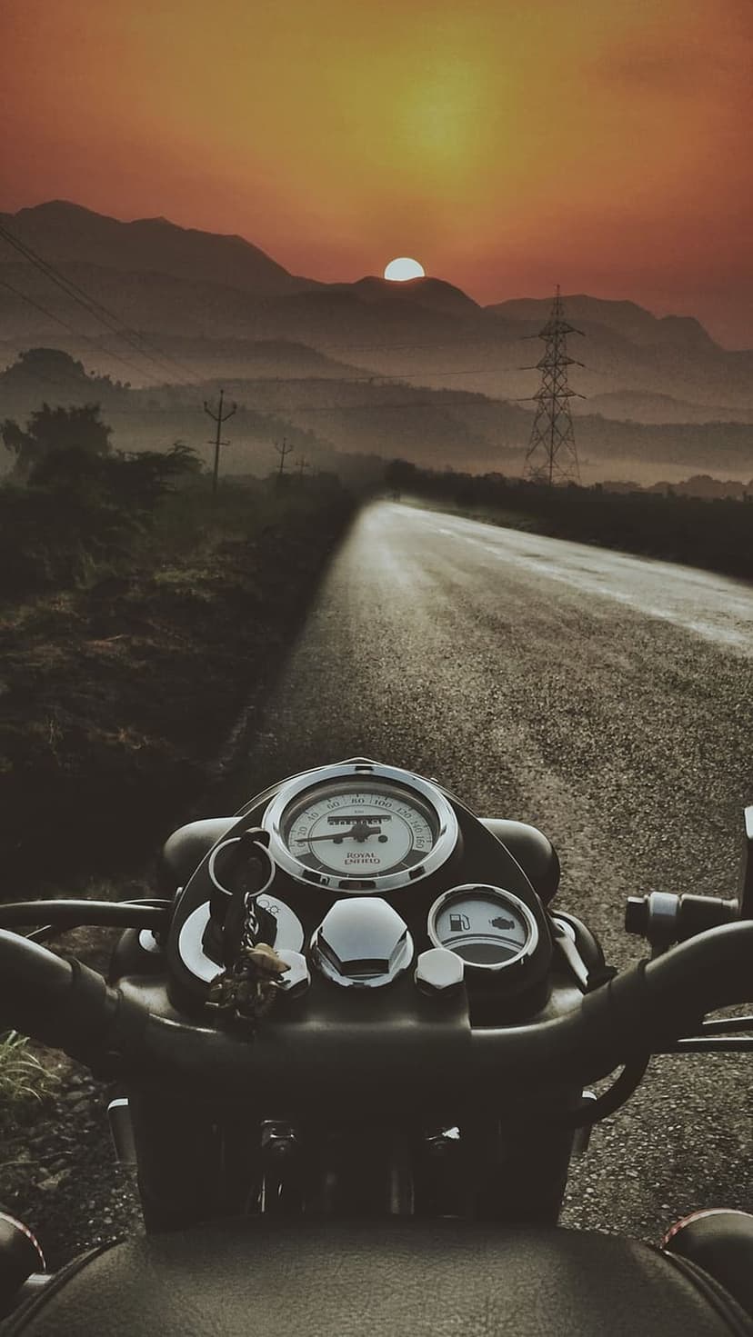 Motorcycle handlebars overlook a winding mountain road during a misty orange sunrise with power lines in view.