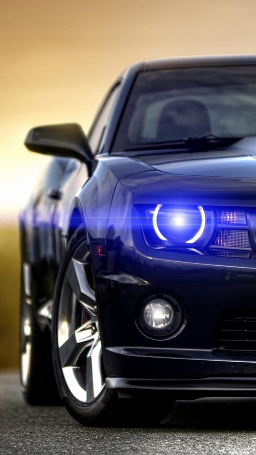 Chevrolet Camaro front grille with glowing blue halo headlights at a low angle against a blurred golden sunset.