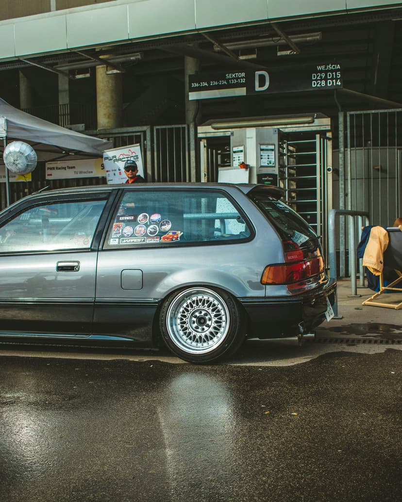 Silver Honda Civic EG hatchback with a lowered stance sits on reflective wet pavement outside a stadium.