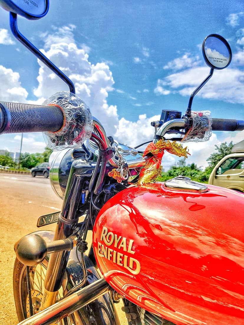 Red Royal Enfield fuel tank with a gold logo reflecting a blue sky and white clouds near chrome handlebars.