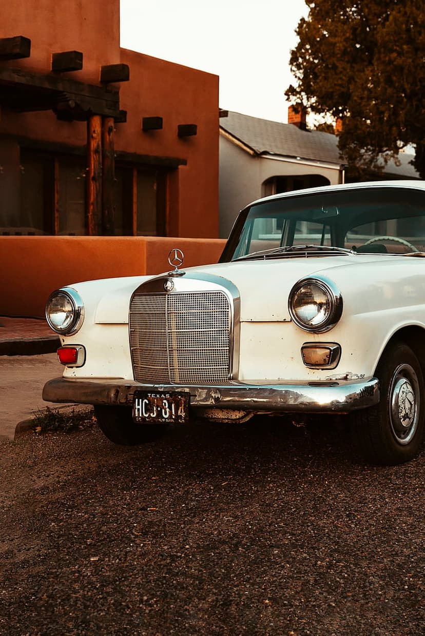 White Mercedes Benz sedan with a chrome grille parked in front of an ochre adobe building during golden hour.