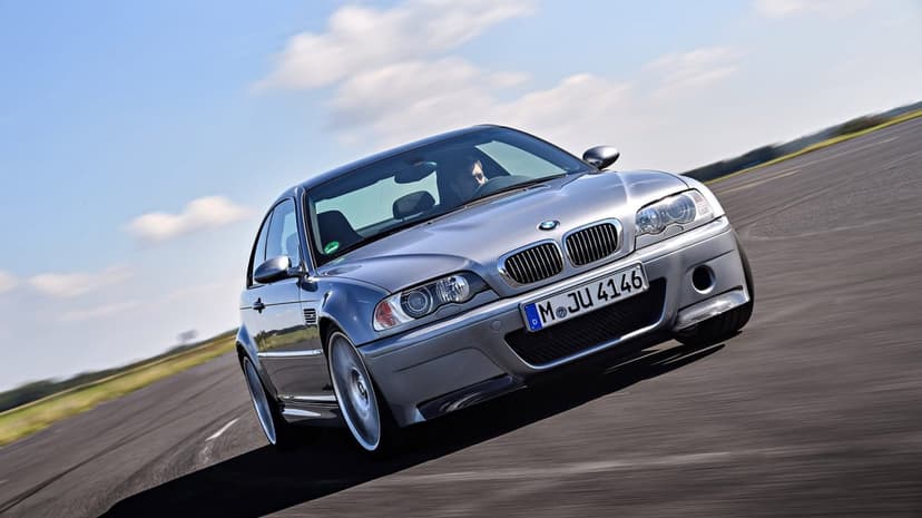 Silver BMW M3 E46 CSL sports car driving fast on an asphalt track under a bright blue sky with light clouds.