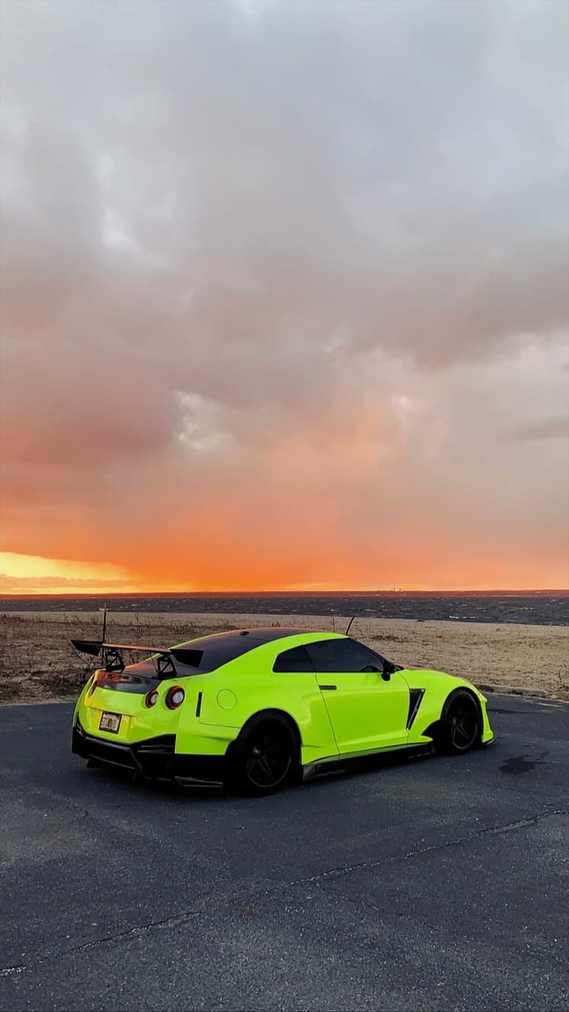 Neon green Nissan GT-R sports car with a wide body kit parked at sunset near a calm ocean and orange sky.