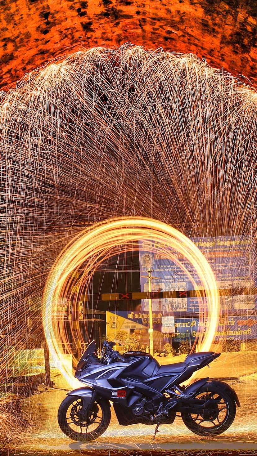 Motorcycle centered in a luminous ring of spinning steel wool sparks against a dark nighttime street scene.