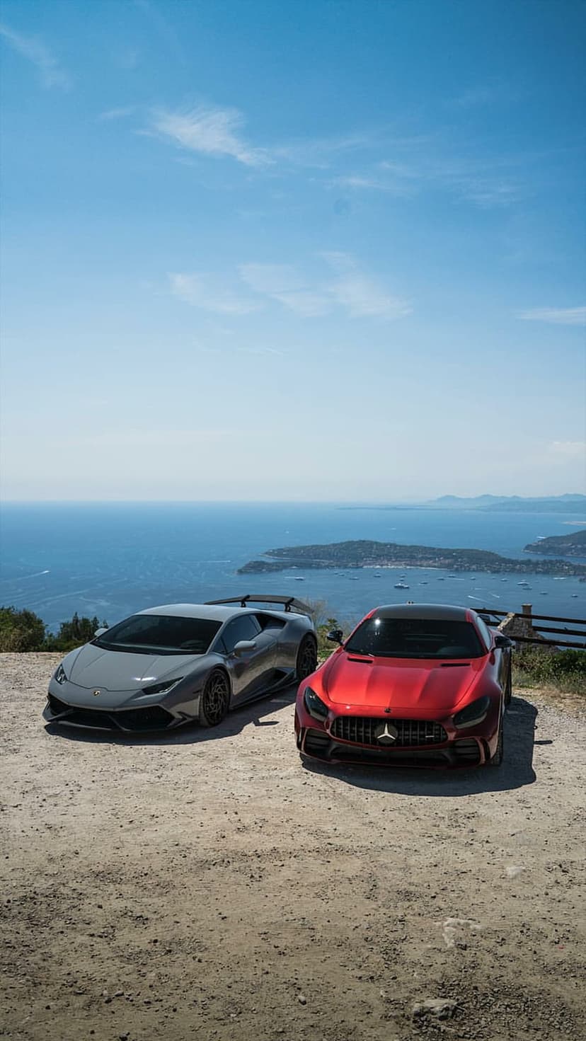 Gray Lamborghini and red Mercedes-AMG GT parked on a dirt cliffside overlooking the blue Mediterranean ocean.