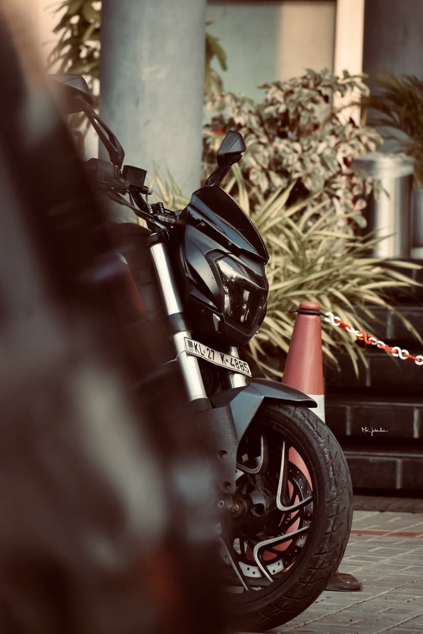 Black motorcycle front wheel and angular LED headlight assembly set against a blurred green foliage backdrop.