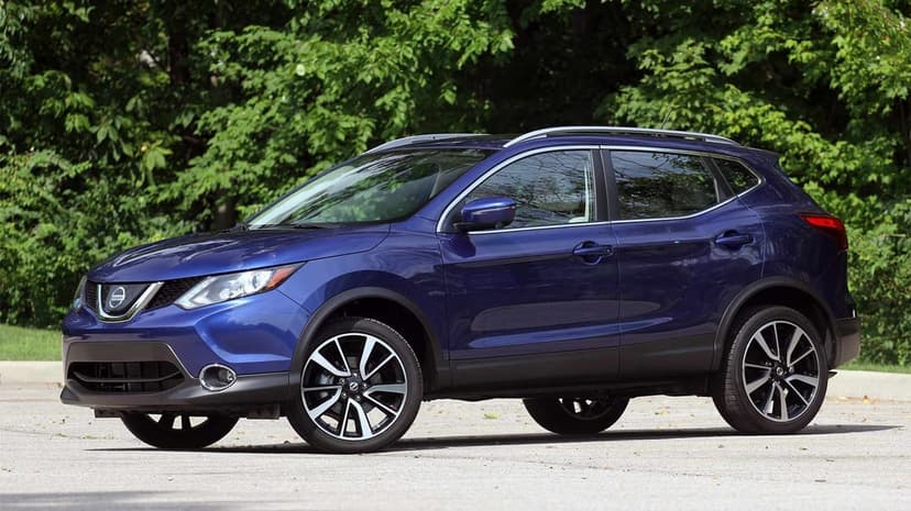 Metallic blue Nissan Rogue Sport parked on asphalt with a backdrop of dense green trees in daylight.