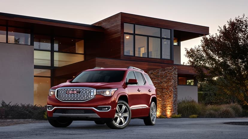 Red GMC Acadia SUV parked in front of a modern wood and glass house at dusk with warm interior lighting.