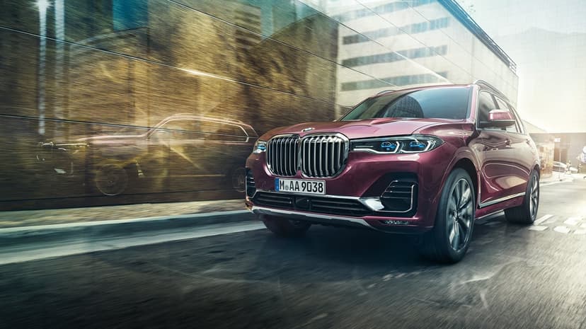 Burgundy luxury SUV driving on a city street with a gold reflection on a glass building facade.