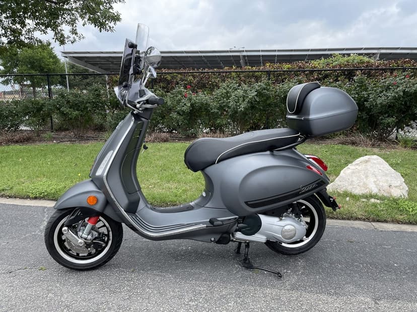 Matte grey Vespa scooter with a windshield and top box parked on pavement next to a green lawn and bushes.